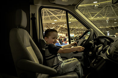 A playful moment of a young child sitting in the driver’s seat of a vehicle, photographed by Justin Adelmann at an indoor event.