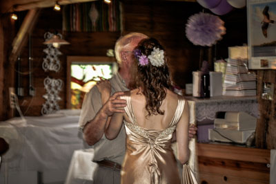 A tender moment of a couple dancing indoors, with the focus on the woman’s elegant satin gown and floral hairpiece, capturing an intimate and nostalgic atmosphere.