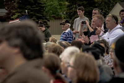 A crowd at an outdoor event, with a man wearing a veteran's hat standing prominently in the middle. The background includes various attendees, trees, and a building.