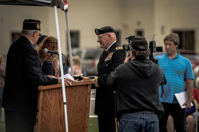 A military ceremony where a uniformed officer shakes hands with a man in a suit. A cameraman is filming the event, and other attendees stand in the background.